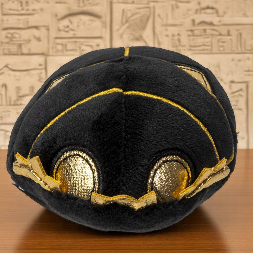Black plush ancient Egyptian Scarab Beetle with gold accents on a wooden surface with an Egyptian-style wall in the background