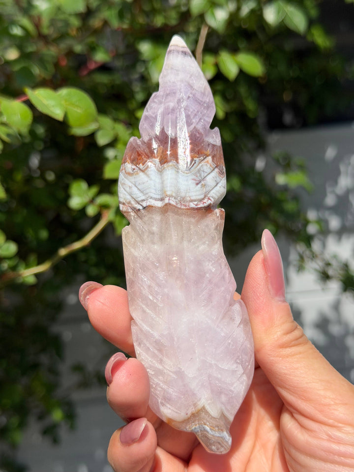 Close-up of a single hand-carved amethyst lace agate feather held in hand outdoors, showing translucent lavender crystal tip and detailed agate banding
