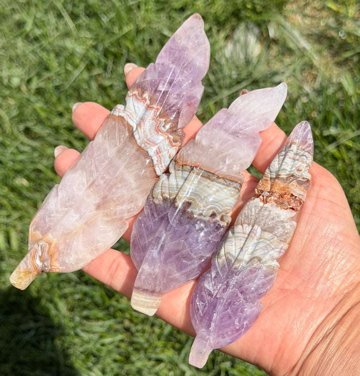 Hand holding three amethyst lace agate feather carvings outdoors, showing size range and purple, lavender, and banded agate color variation