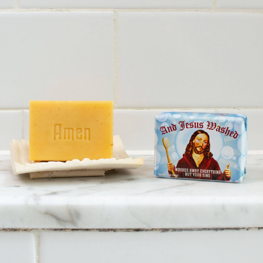 And Jesus Washed almond cocoa butter soap bar embossed with Amen on white soap dish beside novelty gift box on marble counter