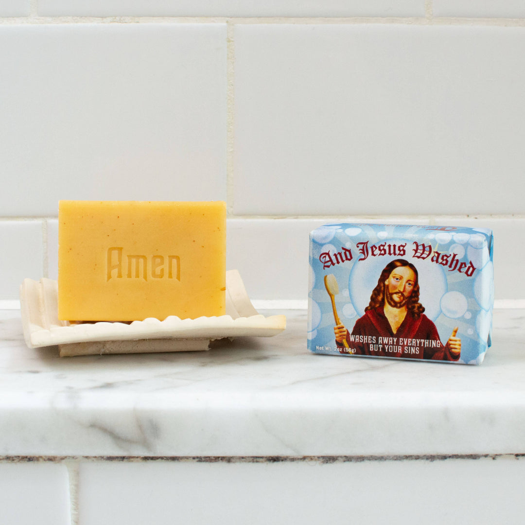 And Jesus Washed almond cocoa butter soap bar embossed with Amen on white soap dish beside novelty gift box on marble counter