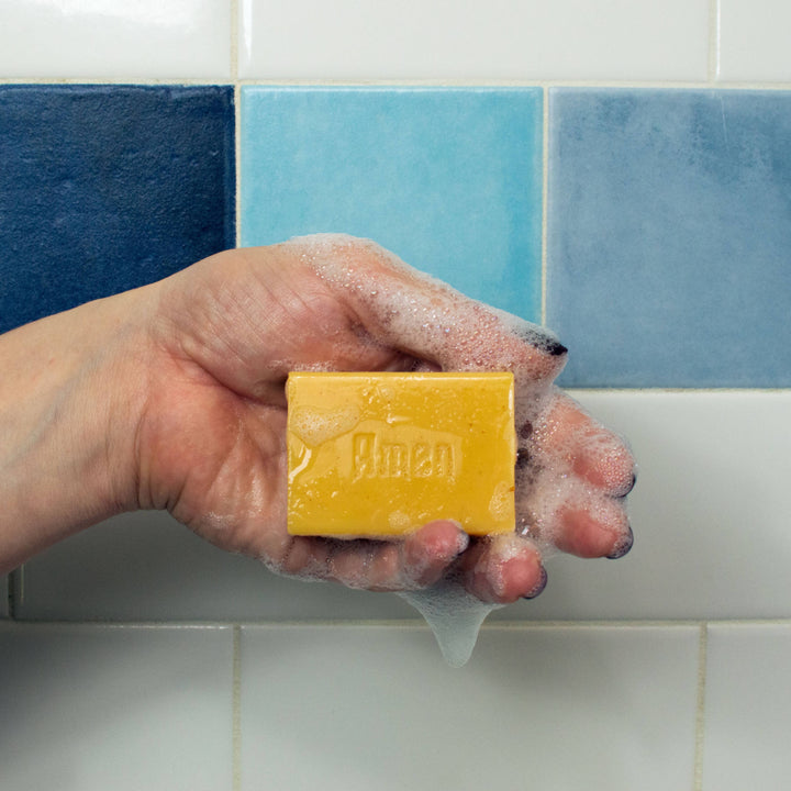Hand holding lathered And Jesus Washed almond cocoa butter soap bar embossed with Amen against blue tile