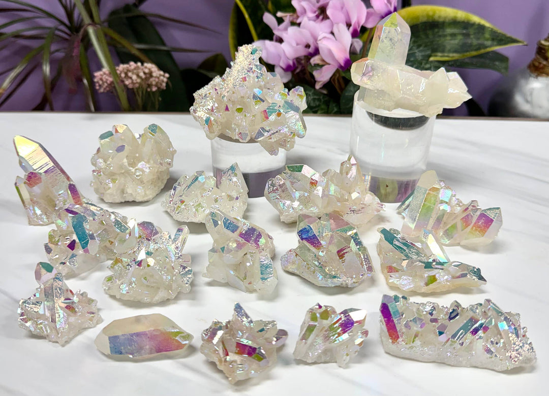 Collection of angel aura quartz clusters in various sizes displayed on a white surface with pink flowers and greenery in the background, showing iridescent rainbow sheen
