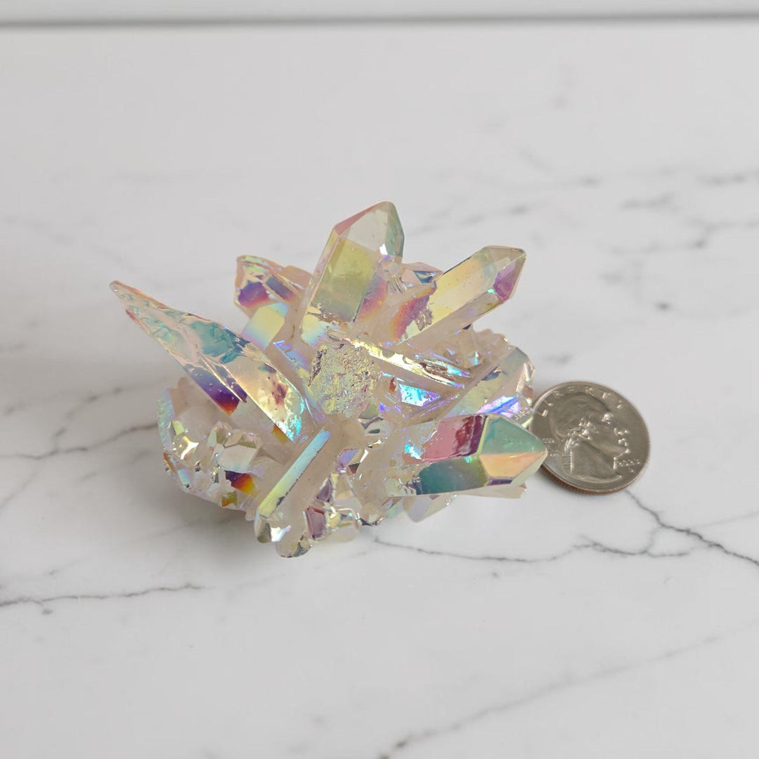 Large angel aura quartz cluster with bright white and blue-green rainbow iridescent sheen displayed next to a quarter for size reference on a white marble surface