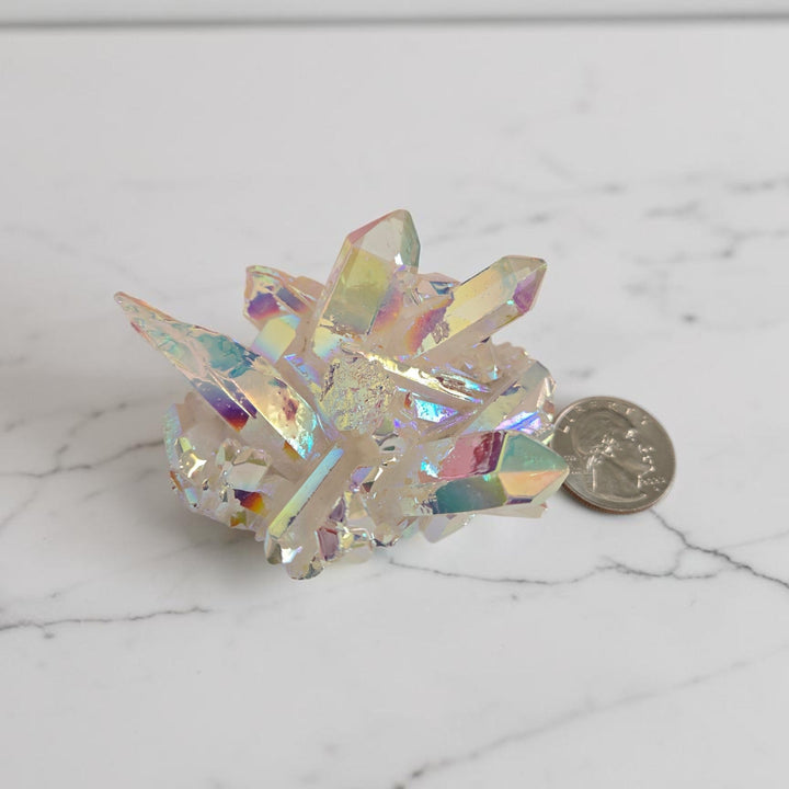 Large angel aura quartz cluster with bright white and blue-green rainbow iridescent sheen displayed next to a quarter for size reference on a white marble surface