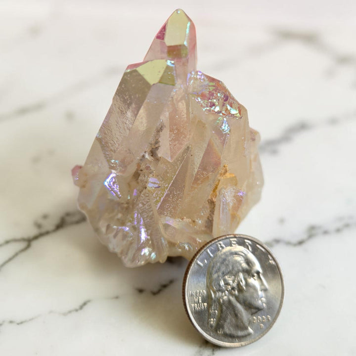 Medium angel aura quartz cluster with soft peachy-pink iridescent sheen displayed next to a quarter for size reference on a white marble surface
