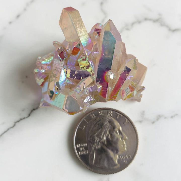 Small angel aura quartz cluster with iridescent pink and rainbow sheen displayed next to a quarter for size reference on a white marble surface