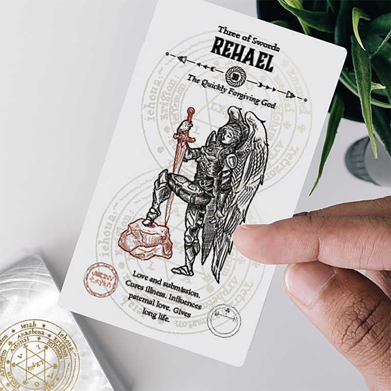 Hand holding the Rehael Three of Swords card from Angel Tarot by Travis McHenry — armored angel with sword, summoning seal visible