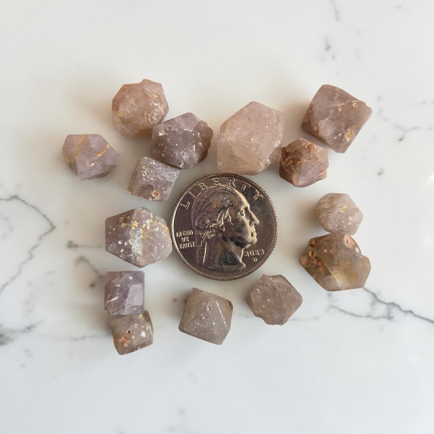 Natural Beta Quartz crystal specimens — 1 oz set of approximately 12 small hexagonal dipyramid pieces in pale mauve-grey with golden inclusions, shown with a US quarter for size reference on white marble