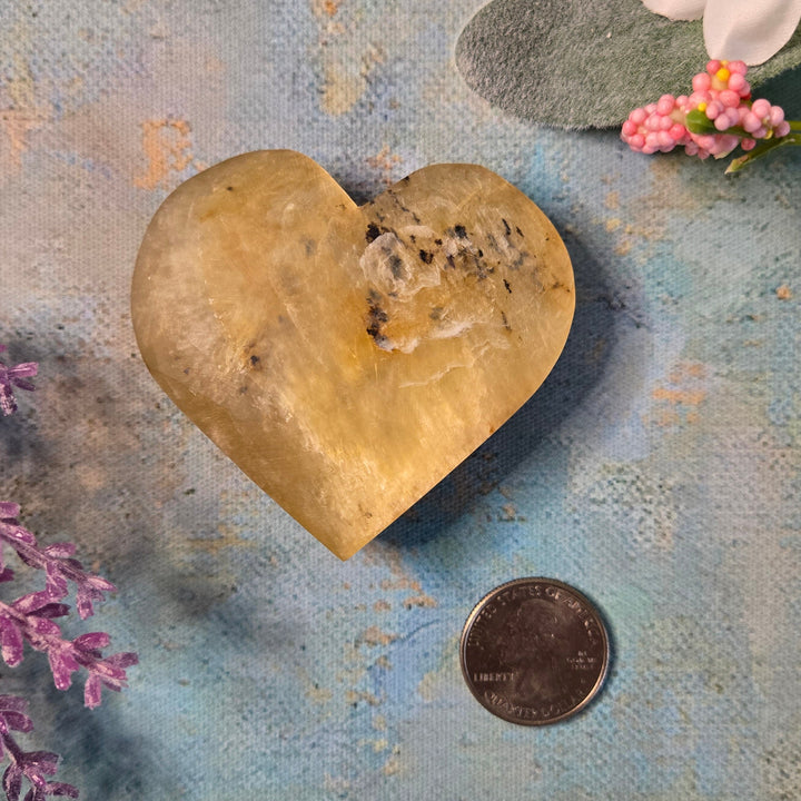 Polished golden mica heart crystal with quarter coin for size reference, approx 2.5 inches, on painted blue surface