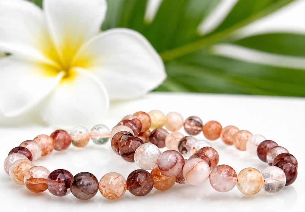 Two natural Hematoid Fire Quartz 8mm stretch bracelets laid flat beside a white plumeria flower