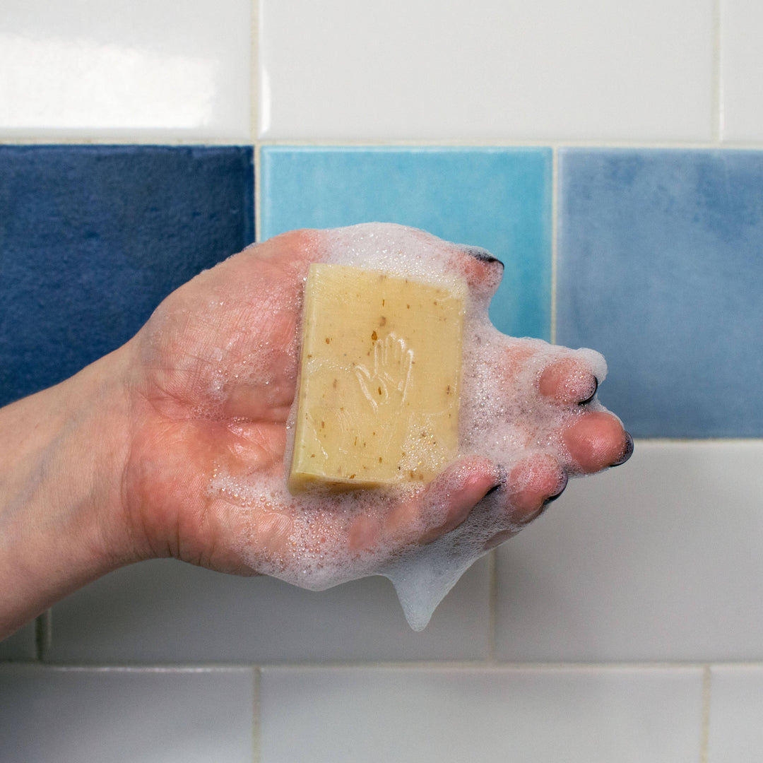 Hand holding lathered palmistry oatmeal exfoliating soap bar with embossed hand imprint against blue tile