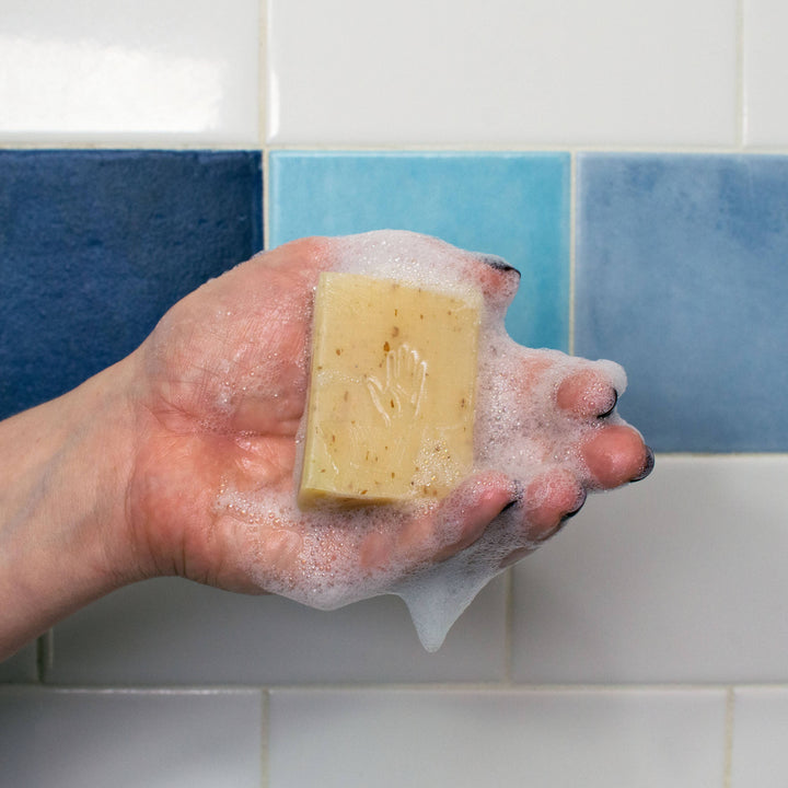 Hand holding lathered palmistry oatmeal exfoliating soap bar with embossed hand imprint against blue tile