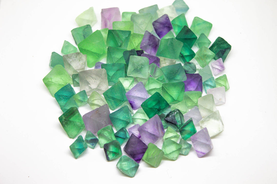 Overhead view of a pile of natural rainbow fluorite octahedrons in green, purple, and clear tones on a white background