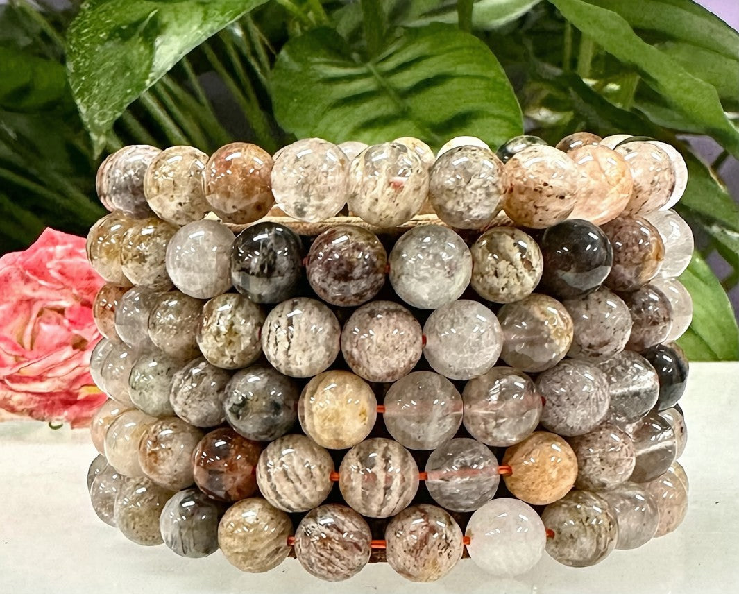 Stack of natural Thousand Layer Phantom Quartz 8mm stretch bracelets showing varied mineral inclusions in earthy tones of grey, brown, and cream, styled with greenery and a pink rose